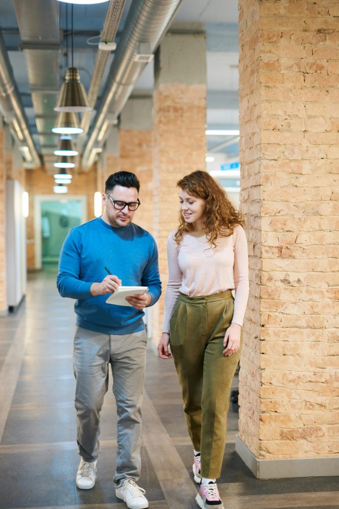 pexels-photo-3182811-3182811 Two business colleagues walking in an office hallway discussing ideas with a notepad.